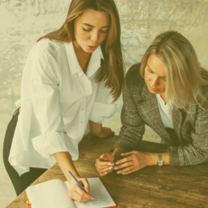 two women leaning on a table looking at a journal with the idea they are coaching or mentoring each other